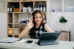 aantrekkelijk gelukkig jong leerling aan het studeren Bij de college bibliotheek, zittend Bij de bureau, gebruik makend van een laptop computer, tablet en hoofdtelefoons hebben een video chatten. foto