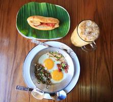 Indo-China gebakken ei met toppings met baguette brood belegd broodje met kaas, ham Aan vers groen banaan blad en ijs koffie Aan houten tafel in eigengemaakt Thais stijl foto