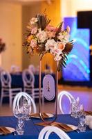 feestelijk tafel met blauw lap, servetten, platen, bestek en bril bereid voor bruiloft gasten in restaurant. mooi bloemen arrangement in goud metaal vaas met aantal vier. verbazingwekkend porties foto