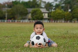 Aziatisch jongen zit met zijn kin Aan een voetbal bal in de gras, in de concept van buitenshuis activiteiten, sport, speeltuinen, vrije tijd activiteiten. zacht en selectief focus. foto