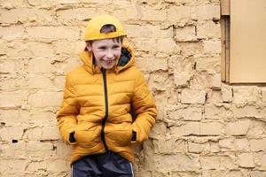 een kind Aan de straat. een gelukkig jongen is staand tegen een steen muur in geel kleren en een basketbal kap. foto
