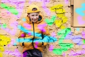 kind in graffitti. de jongen is staand tegen een steen muur in modieus kleren en een basketbal pet, geschetst met helder kleuren. foto