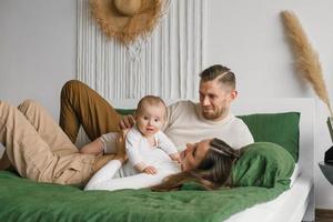 gelukkig jong familie met een baby zoon rust en communiceert met de kind aan het liegen Aan de bed in de slaapkamer foto