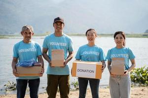 portret team van vrijwilligers vervelend vrijwilliger t-shirt, Holding donaties doos, donaties zak en op zoek Bij camera, staand Aan rivier- achtergrond foto