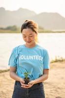 vrouw vrijwilliger Holding pot met groen fabriek glimlachen naar camera staand Aan rivier. bescherming van milieu en natuur, ecologie concept. foto