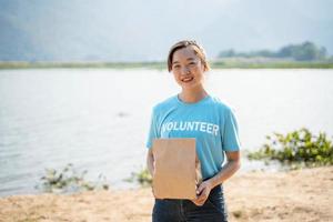 portret van jong vrouw vervelend vrijwilliger t-shirt, Holding bijdrage zak en op zoek Bij camera, staand Aan rivier- achtergrond foto
