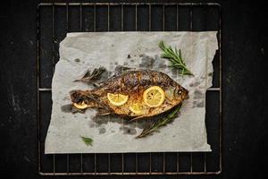 gebakken vis gekookt Aan perkament papier met kruiden, citroen en rozemarijn. barbecue rooster Aan een donker achtergrond. foto