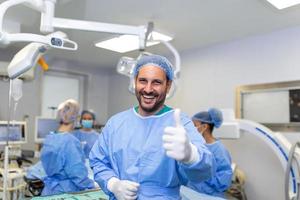 jong mannetje chirurg staand in in werking kamer tonen duimen omhoog, klaar naar werk Aan een geduldig. mannetje medisch arbeider chirurgisch uniform in operatie theater. foto