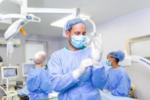 portret van mannetje dokter chirurg zetten Aan medisch handschoenen staand in operatie kamer. chirurg Bij modern in werking kamer foto