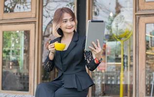 mooi zakenvrouw freelance zittend lang houten bank Holding een koffie kop en gebruik makend van digitaal technologie tablet vdo telefoontje conferentie met klant Bij de koffie winkel buitenshuis terwijl ontspannende gevoel foto