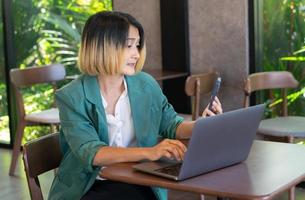zakenvrouw freelance vervelend een pak groen zittend in een cafe en Holding mobiel telefoon video telefoontje met klant en typen Aan een laptop. foto
