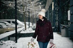 verrukkelijk multi-etnisch vrouw in warm kleren, met boodschappen doen Tassen, wandelen naar beneden de stad straat, Aan een besneeuwd winter nacht foto