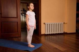 vol lengte portret van aanbiddelijk Kaukasisch weinig meisje oefenen Aan yoga mat, beoefenen yoga Bij knus huiselijk atmosfeer foto