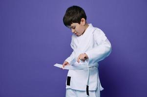 zelfverzekerd Europese tiener- jongen zet Aan wit kimono, banden een riem in de omgeving van zijn taille, klaar voor gevecht sport. aikido vechter. oosters krijgshaftig kunsten foto