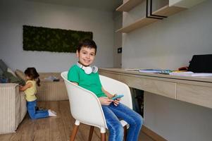 gelukkig tiener- schooljongen zittend Aan de stoel in zijn kamer, met een smartphone in zijn handen, glimlacht met vrolijk glimlach op zoek Bij camera. aanbiddelijk Europese kinderen in kinderen kamer. foto