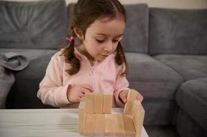 aanbiddelijk Kaukasisch 4 jaren oud baby meisje gebouw huis met houten bakstenen en blokken. hand- beweging controle en concentratie vaardigheden concept, leerzaam bord spellen foto