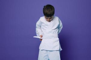 zelfverzekerd Kaukasisch school- leeftijd jongen zet Aan wit kimono, banden een riem in de omgeving van zijn taille, klaar voor gevecht sport. aikido vechter. oosters krijgshaftig kunsten foto