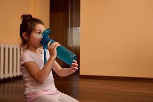 aanbiddelijk kind meisje in wit t-shirt, zittend Aan geschiktheid mat en drinken water van blauw sport- fles foto