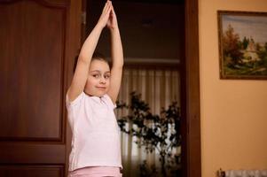 mooi weinig kind meisje verhogen haar armen omhoog, uitrekken haar lichaam, beoefenen yoga, opwarming omhoog terwijl oefenen binnenshuis foto