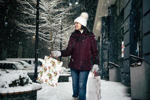 verrukkelijk jong vrouw wandelen naar beneden de straat met boodschappen doen Tassen met Kerstmis geschenk doos in haar handen. vrolijk kerstmis. foto