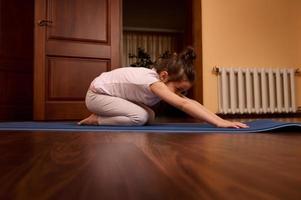 lief baby meisje uitrekken haar lichaam in kind houding, beoefenen yoga Aan een blauw geschiktheid mat in een knus houten huis interieur foto