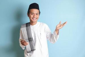 portret van jong Aziatisch moslim Mens Holding mobiel telefoon met glimlachen uitdrukking Aan gezicht terwijl richten vinger naar de kant. geïsoleerd beeld Aan blauw achtergrond foto