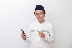 portret van jong opgewonden Aziatisch moslim Mens richten weg met zijn vinger terwijl gebruik makend van mobiel telefoon. geïsoleerd beeld Aan wit achtergrond foto