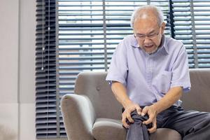 senior Aziatisch Mens lijden van knie artrose symptoom terwijl zittend Aan de bankstel Bij huis met kopiëren ruimte voor medisch chirurgie behandeling en fysiek behandeling concept foto