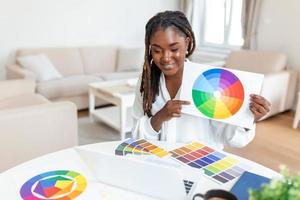 vrolijk Afrikaanse Amerikaans dame ontwerper hebben video conferentie met klanten, zittend Bij bureau in voorkant van computer, Holding kleur paletten, gebaren en lachend, kopiëren ruimte foto