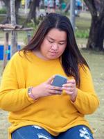 portret dik Aziatisch vrouw lang zwart haar- vervelend geel overhemd zijn gebruik makend van mobiel telefoon of smartphone werk met echt en echt gezicht en uitdrukking binnen park in avond uren zonsondergang dag foto