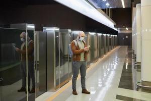 een man met een gezichtsmasker gebruikt een smartphone terwijl hij op een metro wacht
