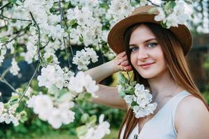 mooi jong meisje in wit jurk en hoed in bloeiend appel boomgaard. bloeiend appel bomen met wit bloemen. foto