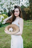mooi jong meisje in wit jurk en hoed in bloeiend appel boomgaard. bloeiend appel bomen met wit bloemen. foto