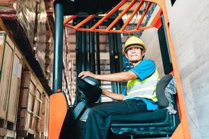 arbeider bestuurder Aziatisch Mens in beschermend veiligheid jumpsuit uniform met geel harde hoed Bij magazijn heftruck lader werken foto