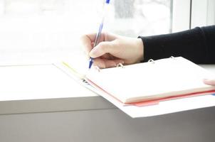 hand- houdt een pen. een persoon schrijft in een notitieboekje in de buurt de venster. foto