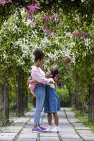 Afrikaanse moeder en dochter knuffelen elk andere terwijl gelukkig wandelen onder de bloesem latwerk boog in de openbaar park gedurende zomer voor familie liefde en zorg in moeder dag viering concept foto