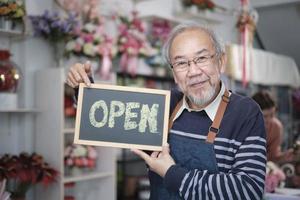 een senior mannetje bloemist eigenaar in schort shows Open teken bord in helder bloem winkel op te slaan met glimlach en looks Bij camera, klein bedrijf opening, bezetting pensioen, gelukkig ouderen mkb ondernemer. foto