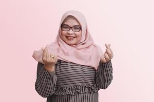 portret van vrolijk Aziatisch vrouw met hijaab, tonen vinger teken van Koreaans liefde. geïsoleerd beeld Aan roze achtergrond foto