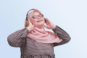portret van vrolijk Aziatisch vrouw met hijaab, genieten van en luisteren naar muziek- gebruik makend van hoofdtelefoon. geïsoleerd beeld Aan blauw achtergrond foto