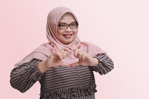 portret van vrolijk Aziatisch vrouw met hijaab, tonen liefde vormig met handen. geïsoleerd beeld Aan roze achtergrond foto