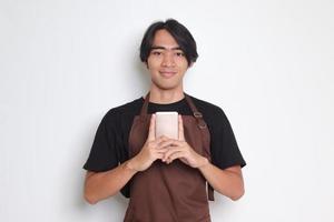 portret van aantrekkelijk Aziatisch barista Mens in bruin schort Holding mobiel telefoon terwijl op zoek Bij camera. geïsoleerd beeld Aan wit achtergrond foto