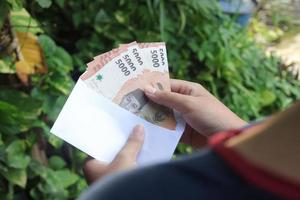 dichtbij omhoog van mannetje hand- invoegen vijf duizend roepia geld bankbiljet in envelop. financieel en spaargeld concept foto