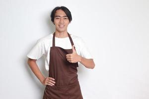 portret van aantrekkelijk Aziatisch barista Mens in bruin schort tonen duim omhoog hand- gebaar. reclame concept. geïsoleerd beeld Aan wit achtergrond foto