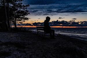 kleurrijk zonsondergang Aan de zee opgemerkt door een Mens in een pet zittend Aan een zwart bank Aan een duin foto