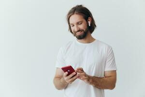 een Mens met een baard in wit t-shirt en jeans looks Bij zijn telefoon flippen door een online sociaal media voeden met hoofdtelefoons in zijn oren luisteren naar een stem bericht Aan een wit achtergrond en glimlachen foto