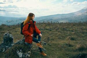 vrouw reiziger in een jasje zit Aan de groot steen en looks Bij de bergen landschap foto