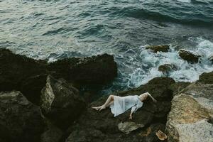 aantrekkelijk vrouw met lang haar- aan het liegen Aan rotsachtig kust met scheuren Aan rotsachtig oppervlakte visie van bovenstaand foto