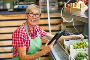 senior vrouw werken in fruit en groenten winkel. ze is Holding groen appels een tablet apparaat. foto