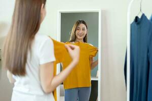 keuze van kleren, niets naar dragen. aantrekkelijk Aziatisch jong vrouw, meisje op zoek in spiegel, proberen Aan verschijnen, kiezen jurk, kleding Aan hanger in garderobe Bij huis. beslissen blouse wat naar zetten Aan welke een foto