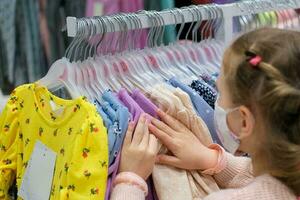 een weinig meisje in een medisch masker kiest een jurk in de op te slaan. een kind is boodschappen doen Bij een kleding winkelcentrum te midden van de covid-19 pandemie. foto